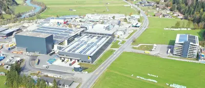 Luftaufnahme eines großen Industriegebäudes mit mehreren angrenzenden Gebäuden und Straßen. Im Hintergrund sind grüne Felder und ein Fluss zu sehen.