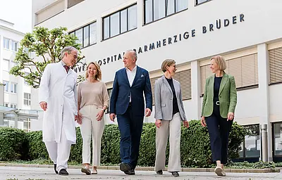 : Fünf Personen gehen gemeinsam zu Fuß vor einem Klinikgebäude. Im Hintergrund ist die Fassade mit Fenstern und der Schrift „Krankenhaus Barmherzige Brüder“ zu sehen. Entlang des Weges befinden sich Grünflächen und ein Baum.