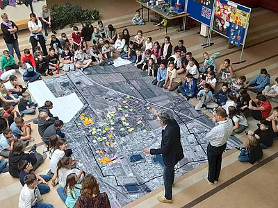 Große Gruppe Kinder sitzt am Boden rundum ein großes Luftbild der Gemeinde. Zwei Erwachsene stehen darauf und leiten an. Post-its kleben am Luftbild