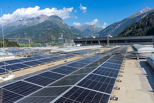 Große Fläche von Solarpaneelen auf einem Flachdach, im Hintergrund eine Berglandschaft unter blauem Himmel mit vereinzelten Wolken.