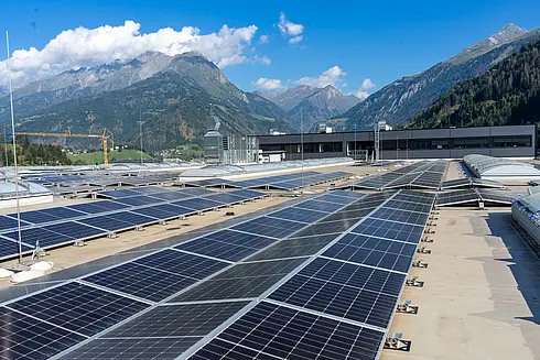 Große Fläche von Solarpaneelen auf einem Flachdach, im Hintergrund eine Berglandschaft unter blauem Himmel mit vereinzelten Wolken.