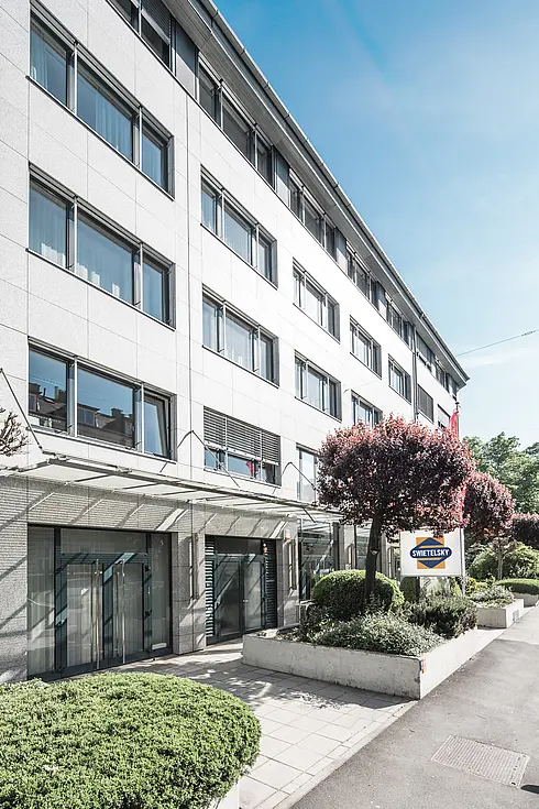 Das moderne Bürogebäude der Swietelsky-Zentrale in Linz. Die Fassade besteht aus grauem Stein mit großen Fenstern, umgeben von gepflegten Grünflächen und Bäumen.