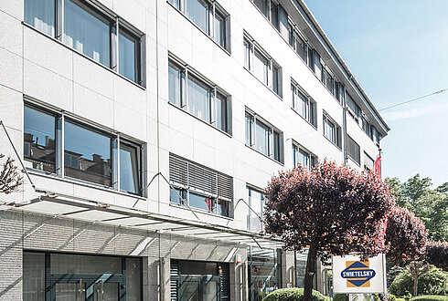 Das moderne Bürogebäude der Swietelsky-Zentrale in Linz. Die Fassade besteht aus grauem Stein mit großen Fenstern, umgeben von gepflegten Grünflächen und Bäumen.