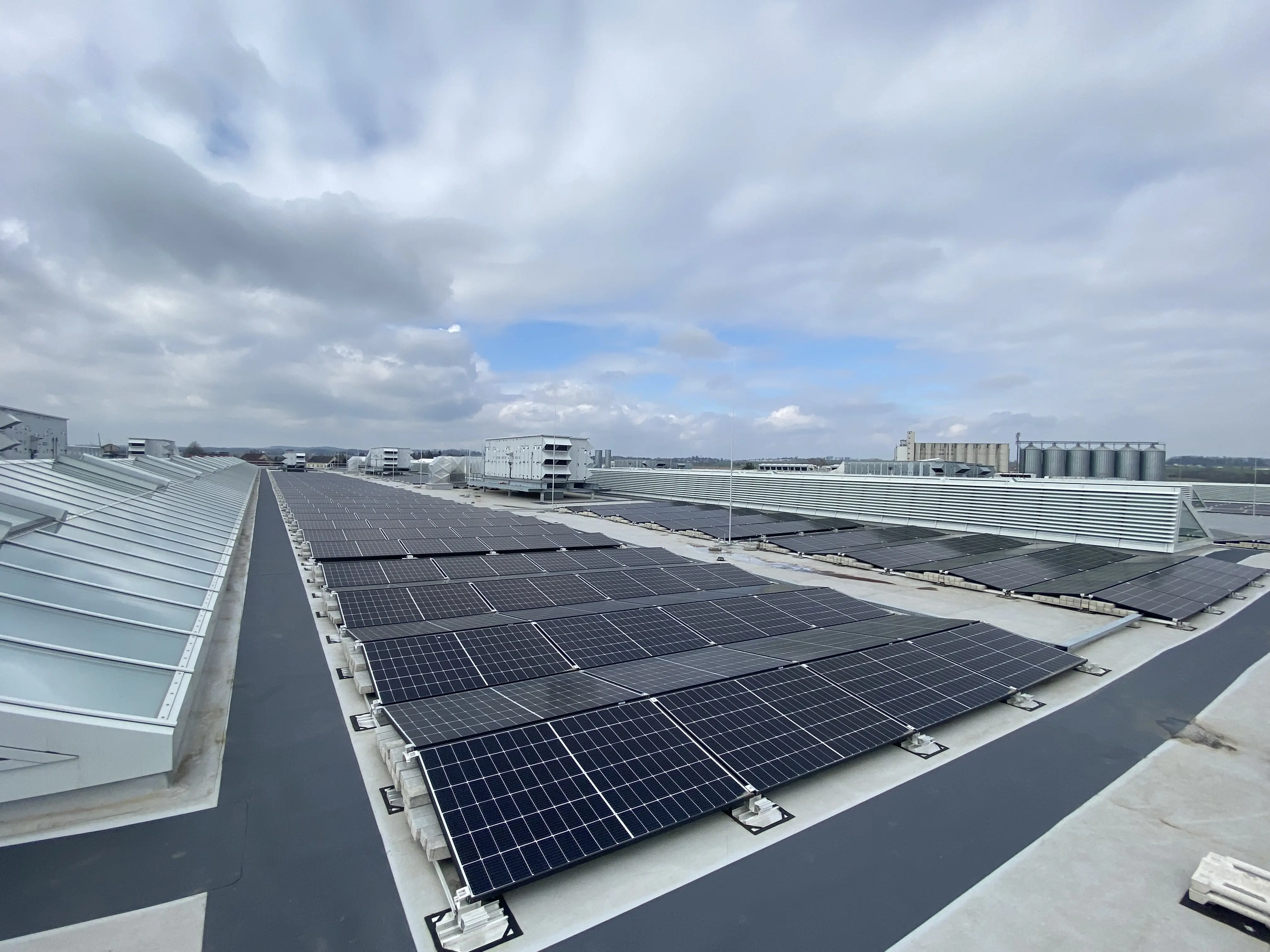 Reihen von Solarmodulen auf einem großen Flachdach unter bewölktem Himmel, mit Blick auf angrenzende Industrieanlagen.