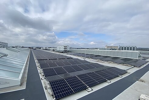 Reihen von Solarmodulen auf einem großen Flachdach unter bewölktem Himmel, mit Blick auf angrenzende Industrieanlagen.