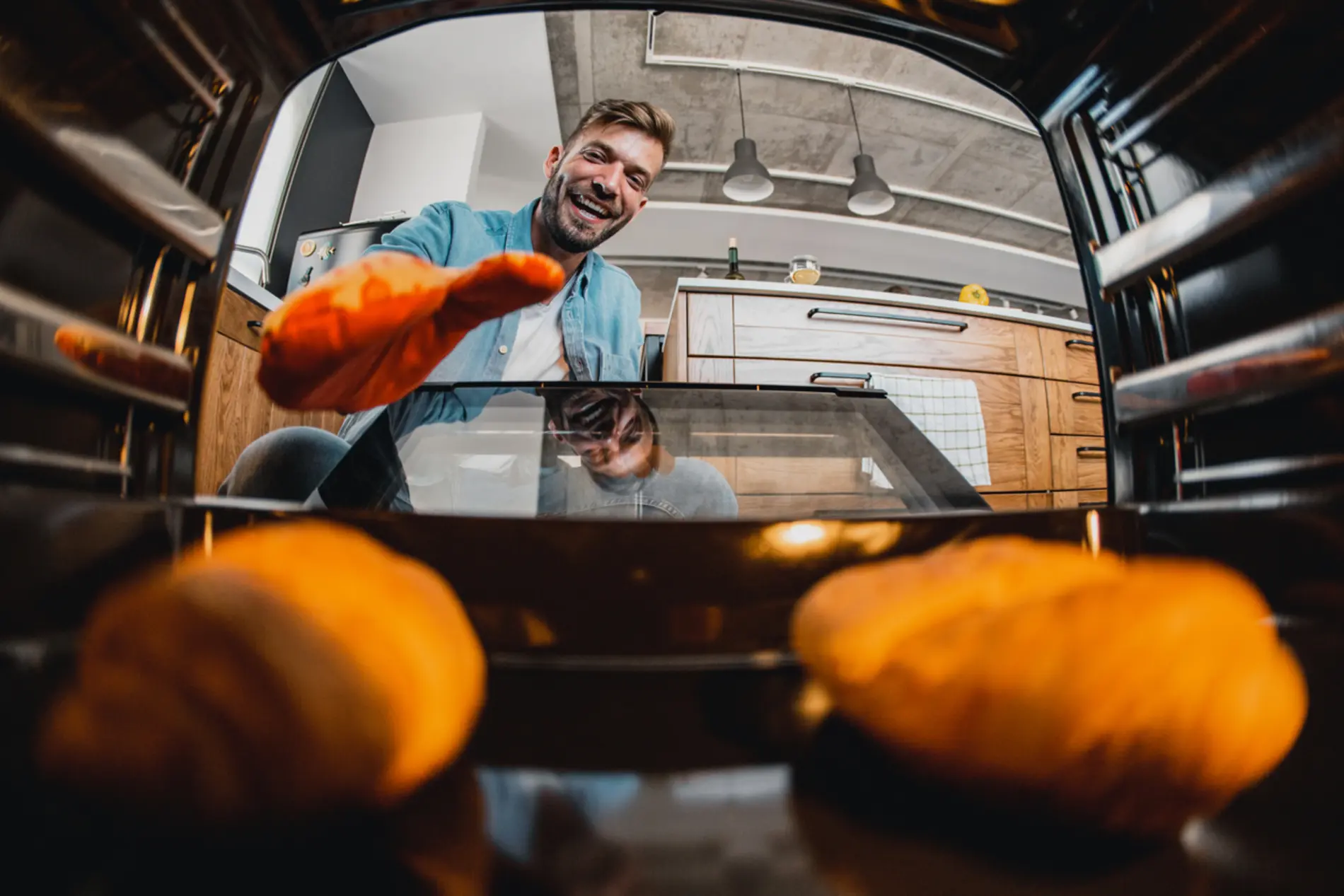 Ein Mann steht freudig vor dem Backofen und nimmt mit seinem Backhandschuh frische Semmeln hinaus.