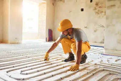 Mann mit Helm und Handschuhen verlegt im Rohbau gerade dünne Rohre für die Bodenheizung