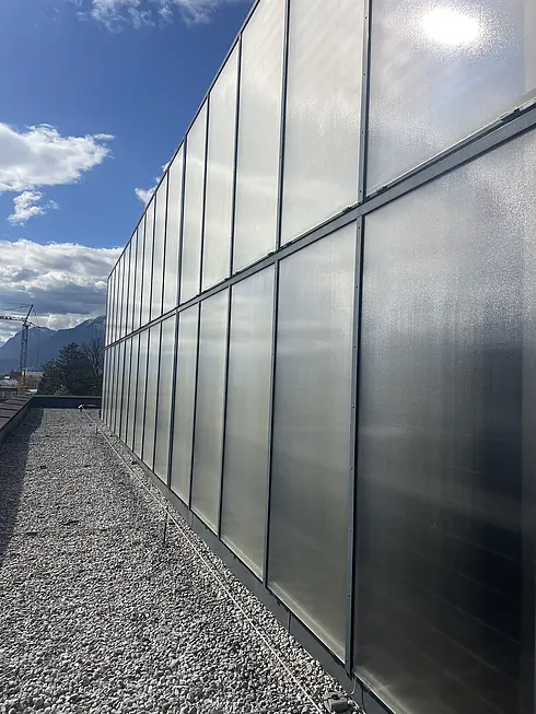 Große, reflektierende Wand mit Gittermuster neben einem Kiesboden. Im Hintergrund sind Berge, Bäume und ein teils bewölkter Himmel zu sehen.