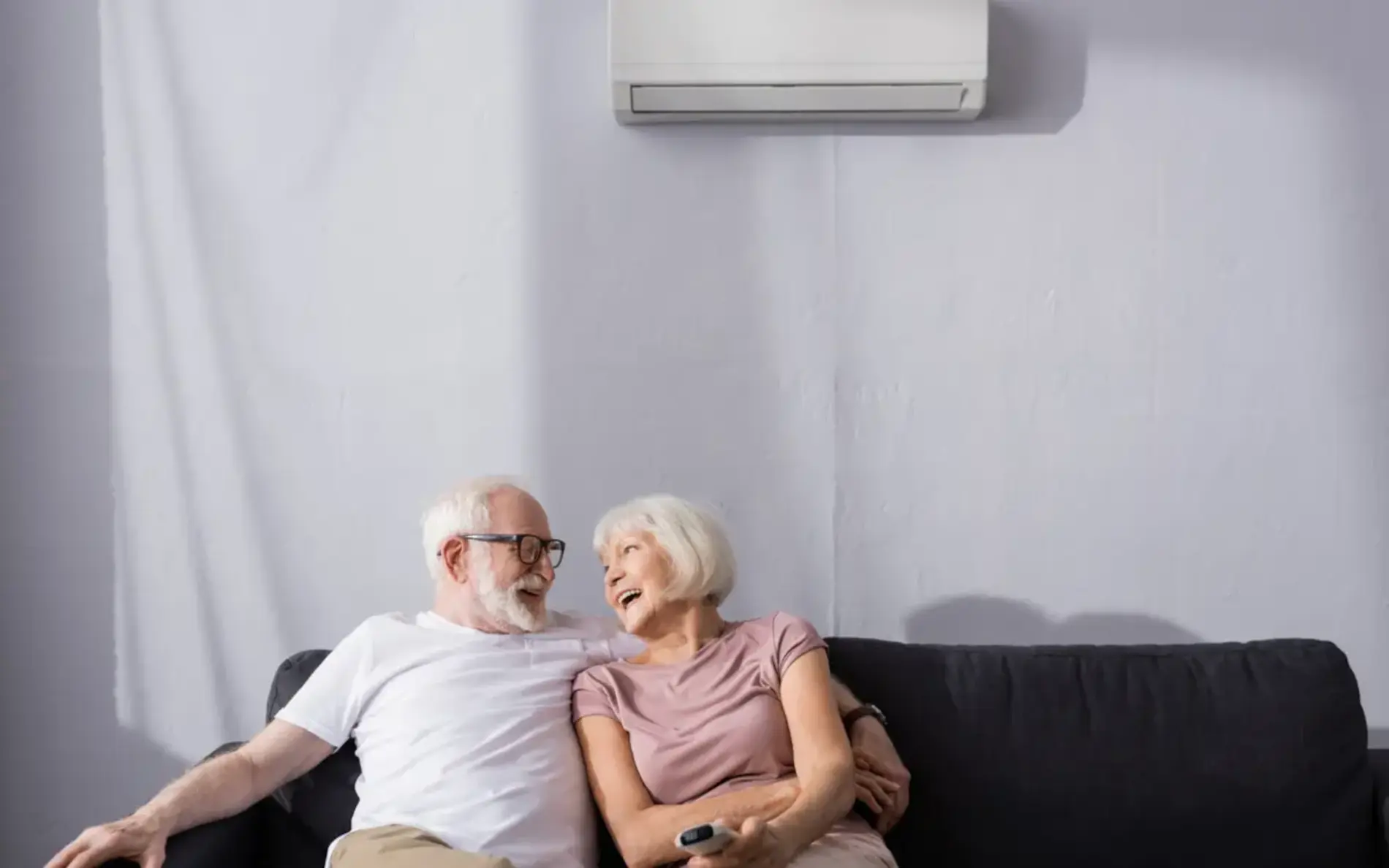 Ein älteres Paar sitzt auf einem Sofa uunter einer Klimaanlage. Der Mann nimmt dabei seine Frau in den Arm während diese ihn anlächelt. 