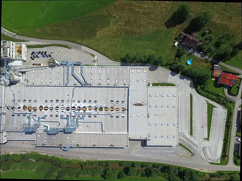 Draufsicht auf ein großes Fabrikdach mit Lüftungsanlagen und Rohrleitungen, umgeben von Parkflächen und Grünflächen.