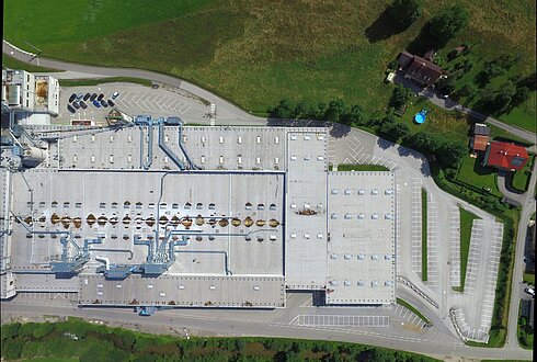 Draufsicht auf ein großes Fabrikdach mit Lüftungsanlagen und Rohrleitungen, umgeben von Parkflächen und Grünflächen.