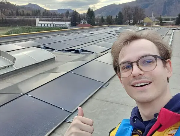Dach mit Solarmodulen und Lichtkuppeln, im Vordergrund eine Person zeigt Daumen hoch, im Hintergrund Berge und Bäume.
