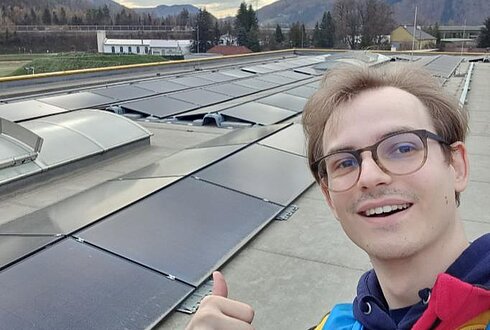 Dach mit Solarmodulen und Lichtkuppeln, im Vordergrund eine Person zeigt Daumen hoch, im Hintergrund Berge und Bäume.