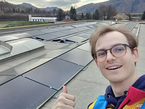 Dach mit Solarmodulen und Lichtkuppeln, im Vordergrund eine Person zeigt Daumen hoch, im Hintergrund Berge und Bäume.