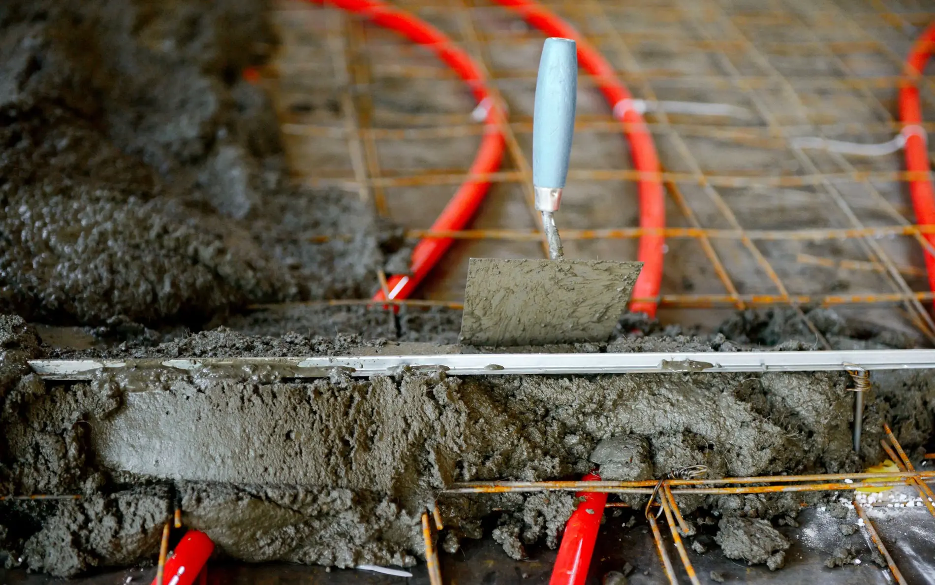Frischer Estrich wird über roten Heizungsrohren und Metallgitter verteilt, um eine Fußbodenheizung zu installieren.
