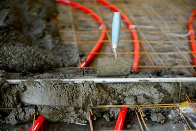Frischer Estrich wird über roten Heizungsrohren und Metallgitter verteilt, um eine Fußbodenheizung zu installieren.