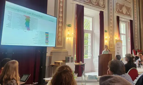 Die Vortragende spricht vor Publikum in barockem Saal. Ein Monitor zeigt die aktuelle Folie.