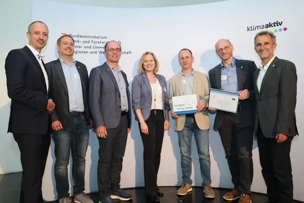 Menschen mit einer Urkunde in der Hand vor einer Fotowand. Sie lächeln in die Kamera.