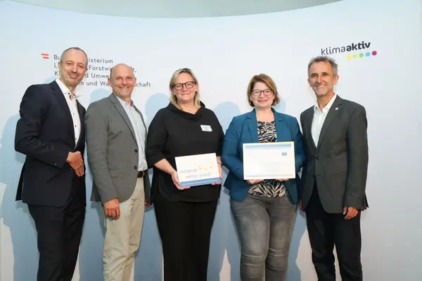 Menschen mit einer Urkunde in der Hand vor einer Fotowand. Sie lächeln in die Kamera.