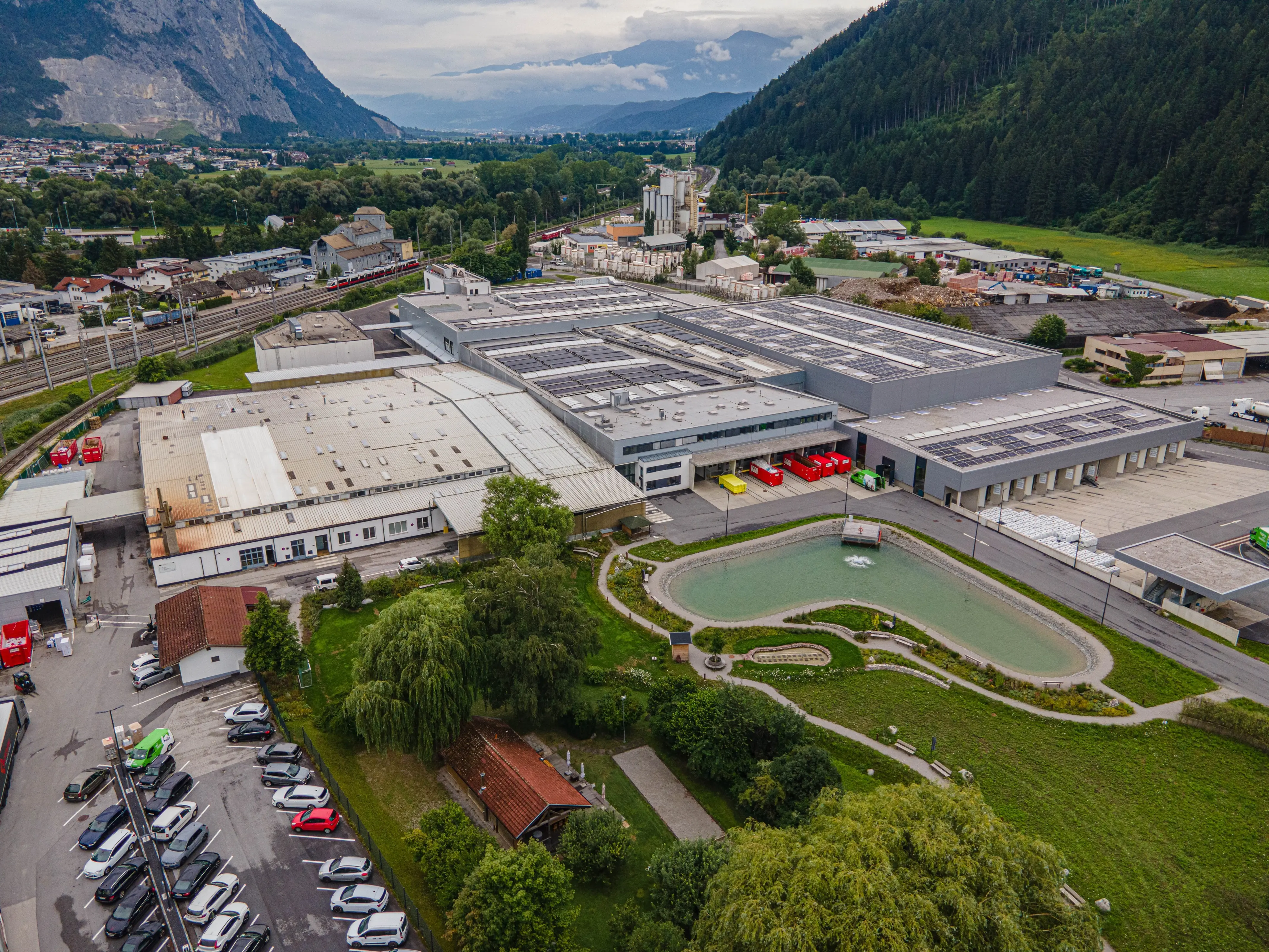 Luftaufnahme eines großen Industrieareals mit Produktionshallen, Parkplätzen und Grünflächen, umgeben von Bergen.