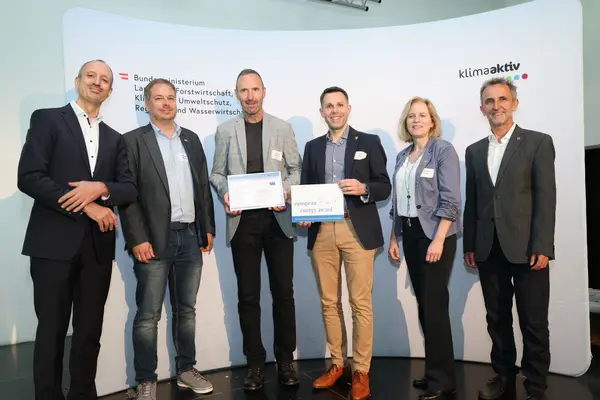 Menschen mit einer Urkunde in der Hand vor einer Fotowand. Sie lächeln in die Kamera.