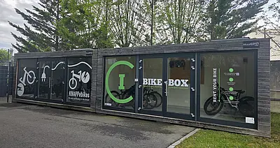 Zwei geschlossene Fahrradgaragen in der Größe von Containern, Glasfront mit Türen, Aufschrift Bike-Box, drinnen Fahrräder, steht im Freien