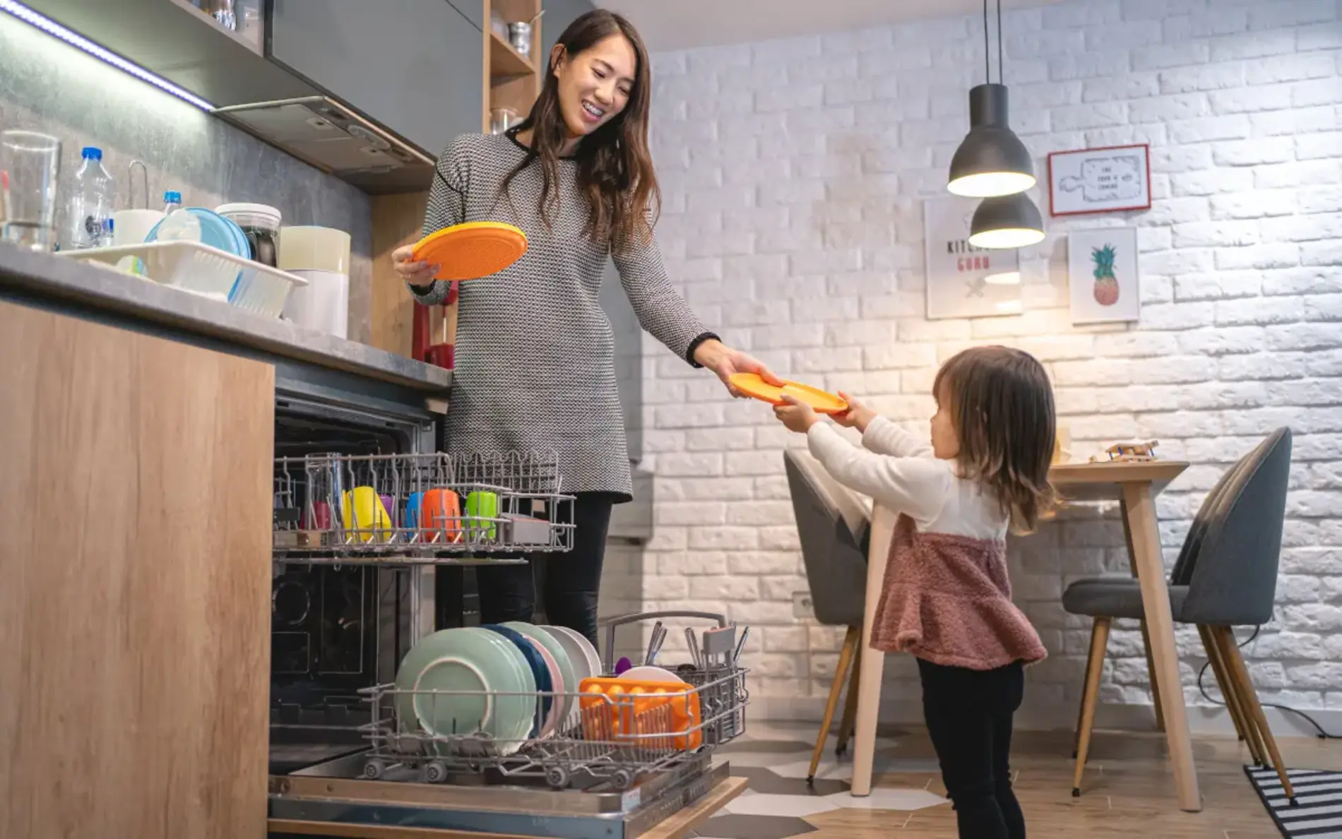 Eine Frau räumt mit einem Mädchen einen Geschirrspüler aus. Sie übergibt dabei ein Telle an das Mädchen.