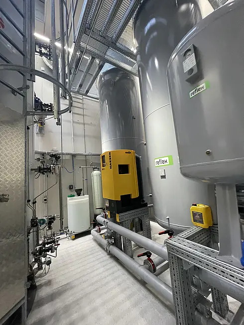 Technikraum mit großen grauen Druckbehältern, gelbem Steuergerät und Leitungen für die Gebäudetechnik.