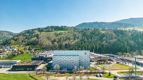 Luftaufnahme eines modernen Gebäudekomplexes mit Glasfassade, umgeben von Wiesen, Bäumen und Hügeln