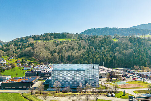 Luftaufnahme eines modernen Gebäudekomplexes mit Glasfassade, umgeben von Wiesen, Bäumen und Hügeln