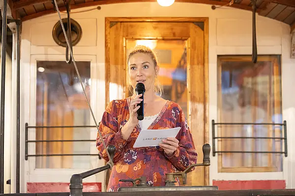Frau spricht ins Mikrofon und hält Moderationskarten. Sie steht auf einer alten Tram, im Hintergrund der Eingang