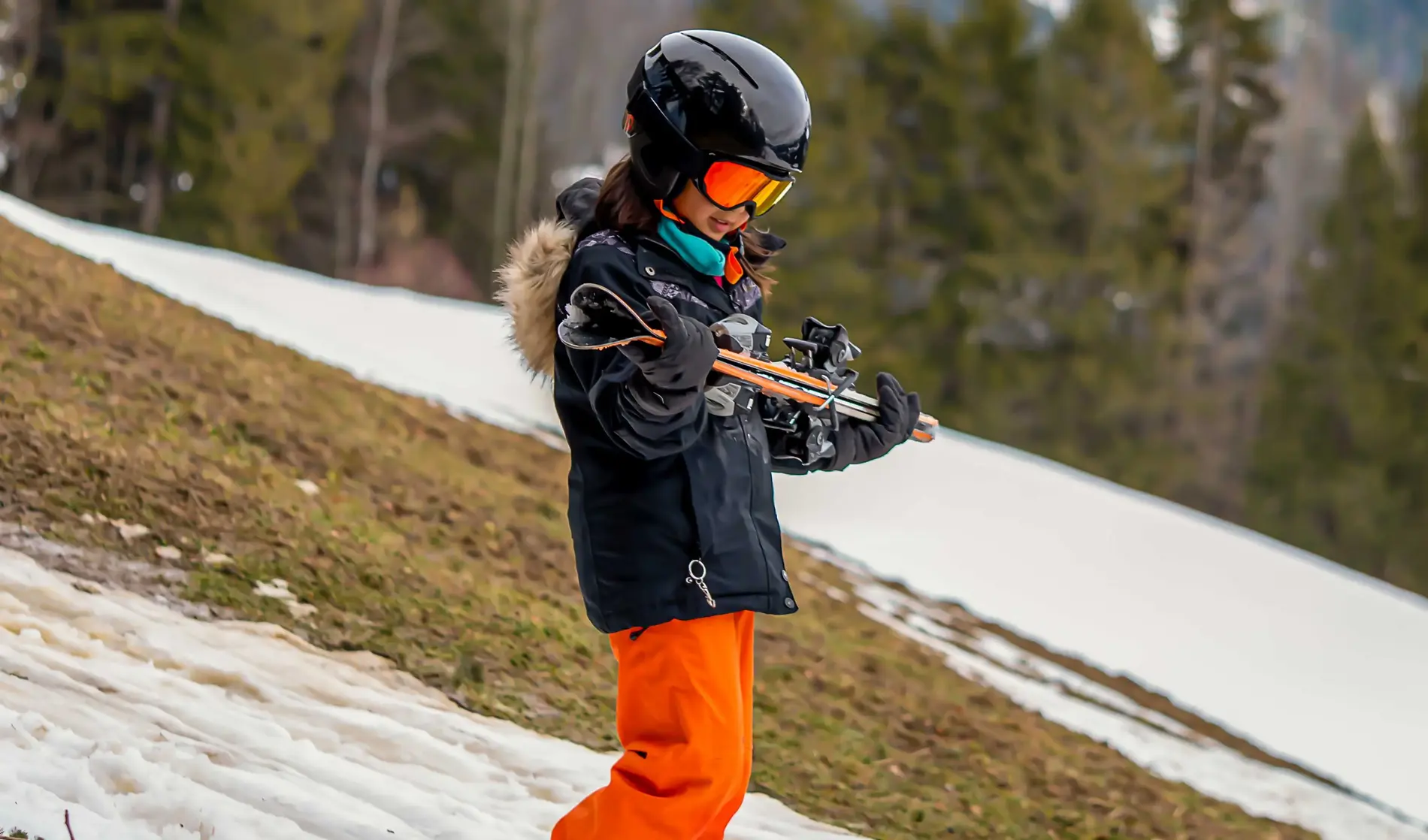 Ein Kind geht im Skianzug mit den Skiern in den Händen an einem kleinen weißen Schneeband hinunter. Im Hintergrund sieht man, dass die Skipiste in weiten Teilen grün ist.