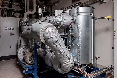 Technische Anlage mit isolierten Rohrleitungen, Pumpen und einem großen zylindrischen Behälter in einem Maschinenraum.