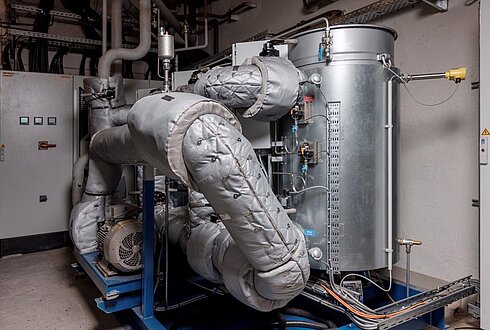 Technische Anlage mit isolierten Rohrleitungen, Pumpen und einem großen zylindrischen Behälter in einem Maschinenraum.