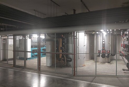 Gitterabgetrennter Bereich mit großen technischen Anlagen, darunter eine Wärmepumpe und Rohrleitungen.