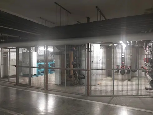 Gitterabgetrennter Bereich mit großen technischen Anlagen, darunter eine Wärmepumpe und Rohrleitungen.