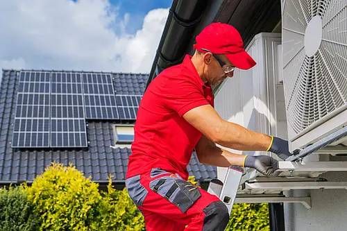 Ein Arbeiter im roten Anzug installiert gerade eine Luftwärmepumpe an der Außenwand eines Hauses. Im Hintergrund ist eine PV-Anlage zu sehen. 