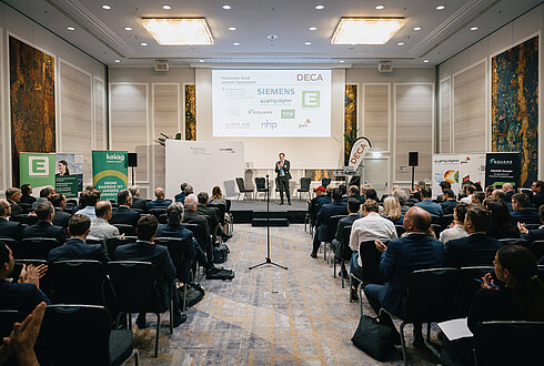 Konferenzszene in einem großen, hellen Raum mit einem Redner auf der Bühne vor einem aufmerksamen Publikum. Im Hintergrund sind ein großer Bildschirm mit Firmenlogos wie DECA, SIEMENS, Energie Steiermark und RHP Technology GmbH sowie mehrere Banner und Poster zu sehen, die verschiedene Unternehmen und Organisationen präsentieren.