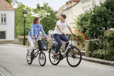 Zwei Personen fahren Fahrrad: Eine Frau mit Faltrad, ein Mann mit Fahrrad. Sie befinden sich in einer typisch österreichischen Gemeinde.
