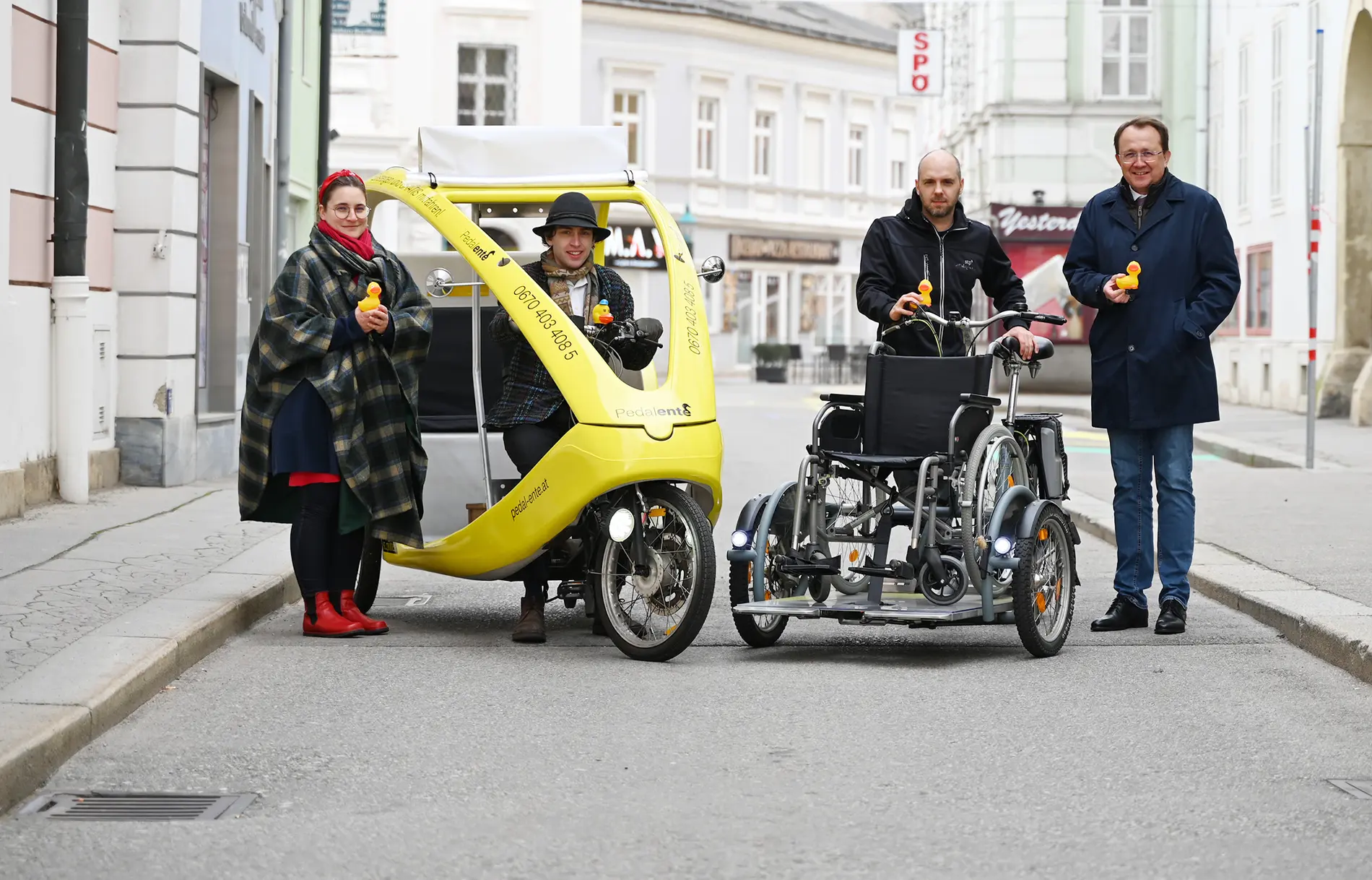 Vier Personen präsentieren in der Innenstadt von St. Pölten ein gelbes Fahrrad-Taxi (die Pedalente) und ein Spezialfahrrad mit Rollstuhlhalterung. Alle halten gelbe Quietscheenten, die für die Pedalente steht.