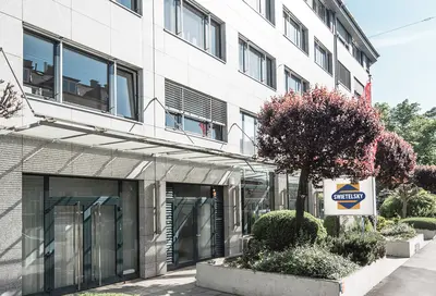 Das moderne Bürogebäude der Swietelsky-Zentrale in Linz. Die Fassade besteht aus grauem Stein mit großen Fenstern, umgeben von gepflegten Grünflächen und Bäumen.