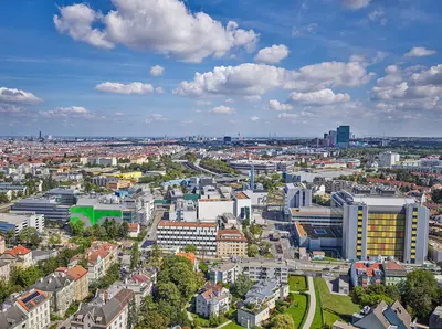 Luftaufnahme eines urbanen Gebiets mit moderner Architektur, Grünflächen und Wohnhäusern unter blauem Himmel mit Wolken.