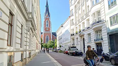 Fahrradstraße Argentinierstraße in rot, links davon ein Gehweg, rechts Parkplätze. Im Vordergrund fährt gerade eine Radfahrerin vorbei, im Hintergrund ein Kirchturm.