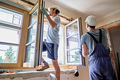 Bei einem offenen Fenster steht ein Bauarbeiter und baut ein Fenster ein. Ein anderer Bauarbeiter ist ihm behilflich. 