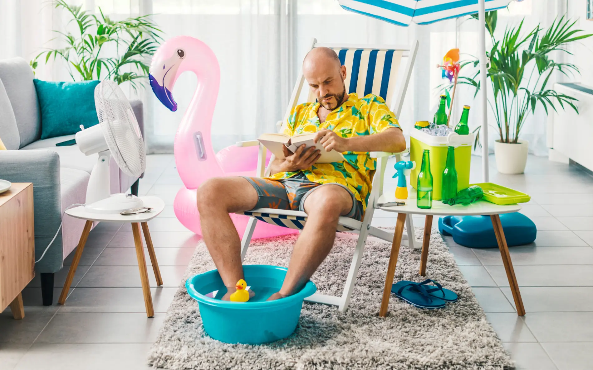Ein mann liegt auf einem Sonnenstuhl im Wohnzimmer, hat ein Hawaiihemd und eine Badehose an und seine Füße kühlt er in einem kleinen Becken ab. Er liest ein Buch. Im Hintegrund ist ein Sonnenschirm und ein aufblasbarer Flamingo abgebildet