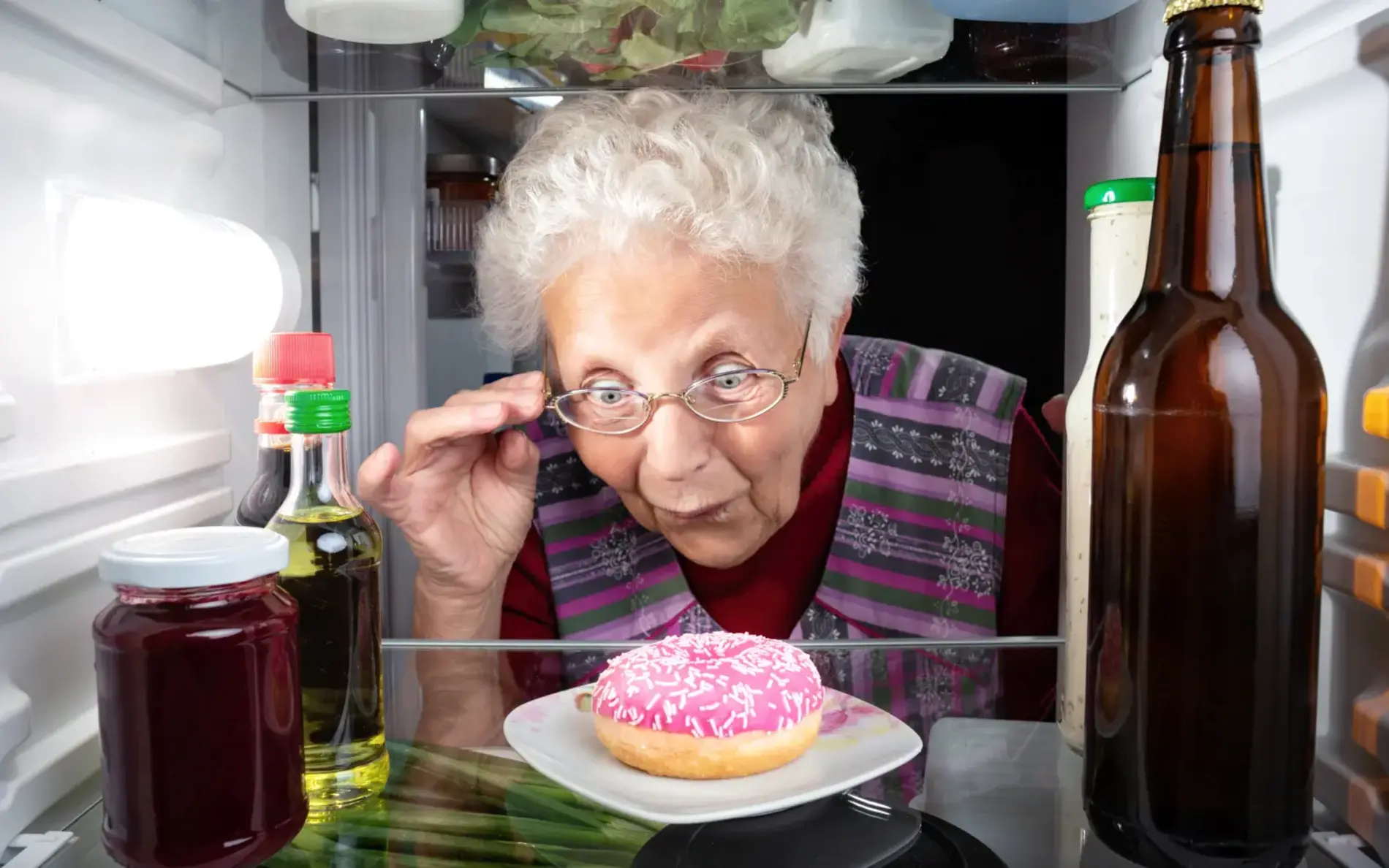 Eine alte Frau steht erstaunt an der Kühlschranktür und schaut erstaunt auf einen pinken Donut