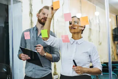Zwei Personen beschriften und kleben Haftnotizen auf eine Glaswand