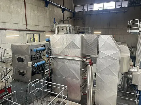 Industrielle Umgebung mit großen metallischen Maschinen und Anlagen. Sichtbar sind verschiedene Rohre, Ventile und Steuerpulte. Die Anlage ist sauber und gut beleuchtet, mit Metallgeländern und Plattformen für den Zugang zu verschiedenen Bereichen der Ausrüstung.