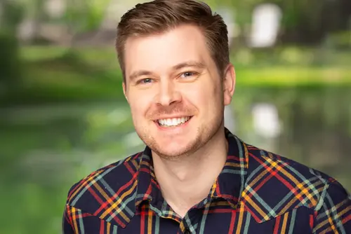 Will Howard is shown outdoors in front of a soft, green, natural background. He is wearing a plaid shirt with dark and bright colored stripes and is photographed from the shoulders upward.