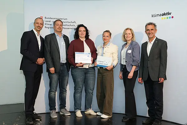 Menschen mit einer Urkunde in der Hand vor einer Fotowand. Sie lächeln in die Kamera.